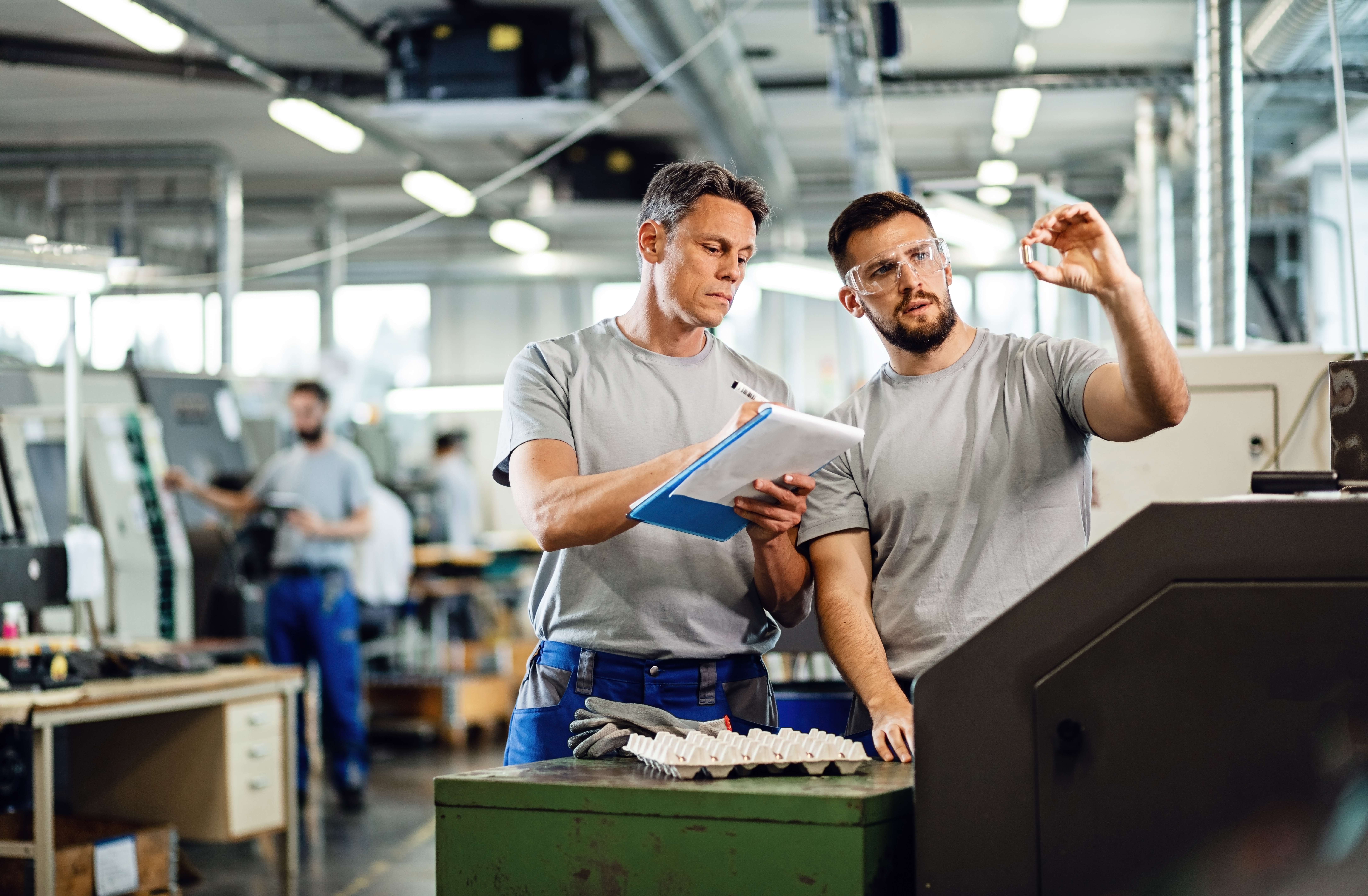 TWO CNC machine operators examining finished products and writing notes in industrial facility.