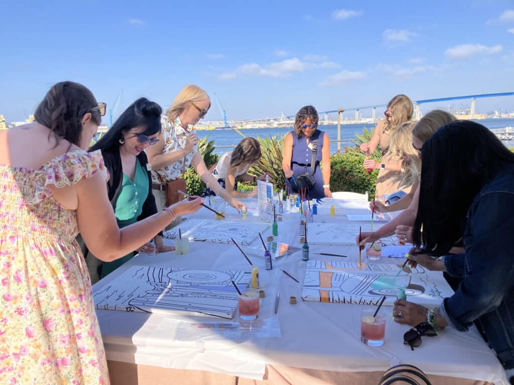 women painting canvases around a table in front of San Diego Bay and the 75 bridge